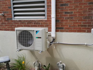 Outdoor wall-mounted air conditioner unit on a brick building above utility pipes and a small plant.