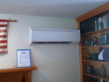 Wall-mounted white air conditioner above shelves and a framed sign in a room.