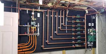 Basement wall with exposed copper plumbing pipes and valves on a dark panel, next to a white door.