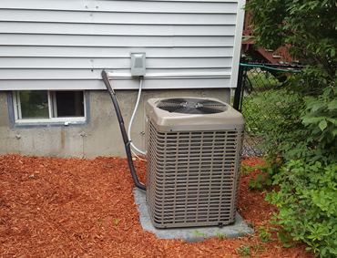 Outdoor air conditioner unit beside a house wall with utility lines and mulch ground cover