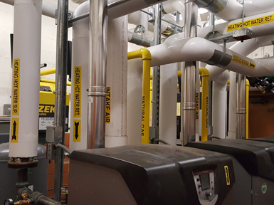 Industrial pipes and valves in a mechanical room, with white, yellow, and black piping overhead.