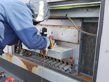 Technician in blue hoodie repairs electrical equipment inside an open metal panel with exposed wiring.