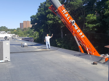 Worker guides an orange crane boom on a flat rooftop beside trees and buildings.