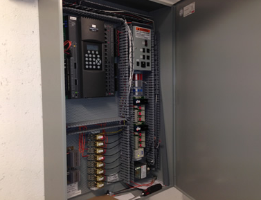 Open electrical control panel with wiring, breakers, and terminal blocks mounted on a wall.