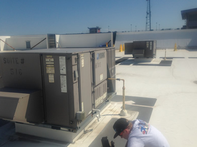 Roof worker beside a gray HVAC unit on a bright, flat rooftop.