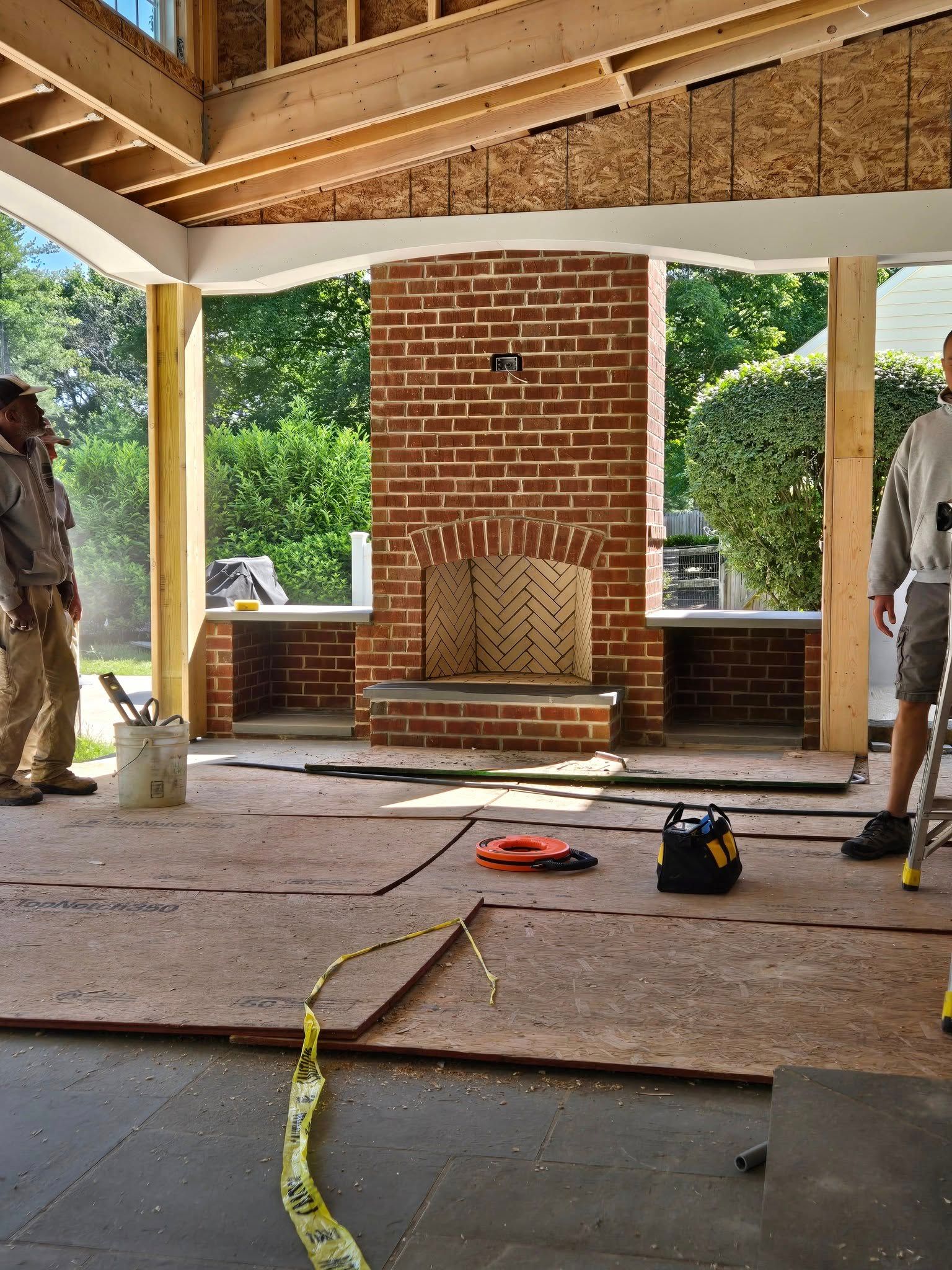 Construction of a brick fireplace on an outdoor porch. Two workers stand nearby the unfinished structure, one wearing a work uniform and the other on a ladder.