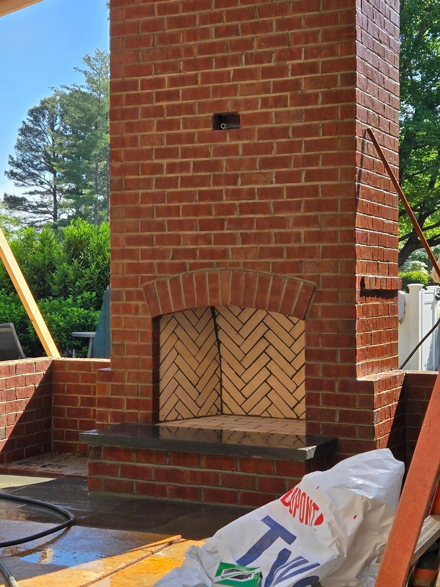 Brick fireplace under construction, featuring a curved opening and herringbone-patterned interior. Red bricks and a blue sky are visible.