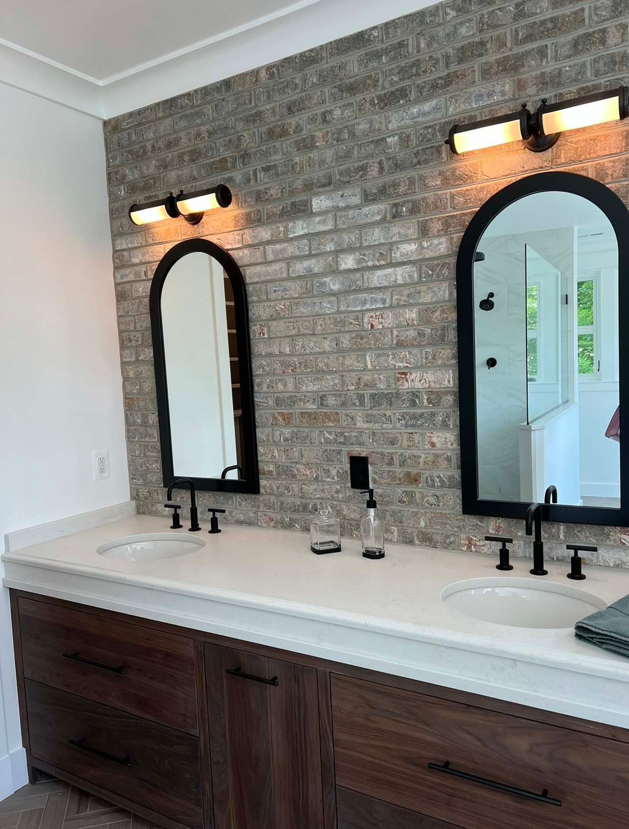 a bathroom with two sinks , two mirrors and a brick wall .