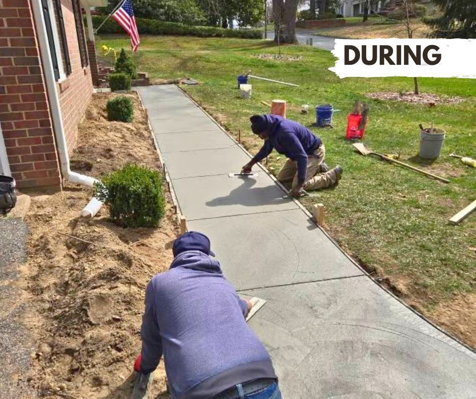 Two workers finishing a concrete sidewalk during construction; one kneels and smooths the surface. The setting is a residential front yard.
