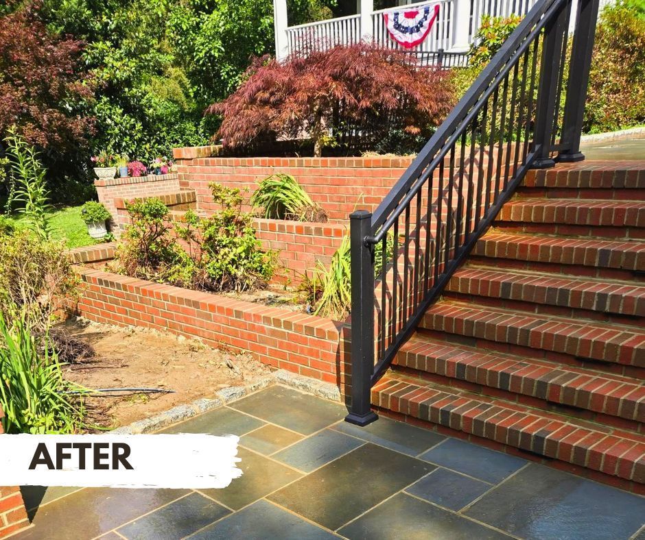 Brick steps and retaining walls lead to a porch with a black railing, flag, and lush landscaping.