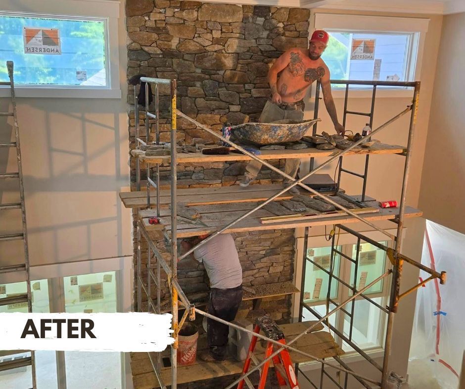 Two workers building a stone fireplace, using scaffolding indoors. One is working from a higher level with a bucket of mortar; the other is working from a lower level.