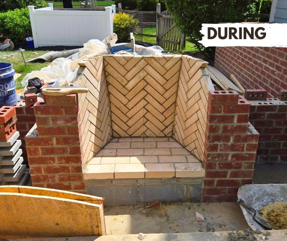 Brick fireplace under construction, featuring herringbone pattern interior. Red bricks, gray cement blocks, and outdoor setting with greenery.
