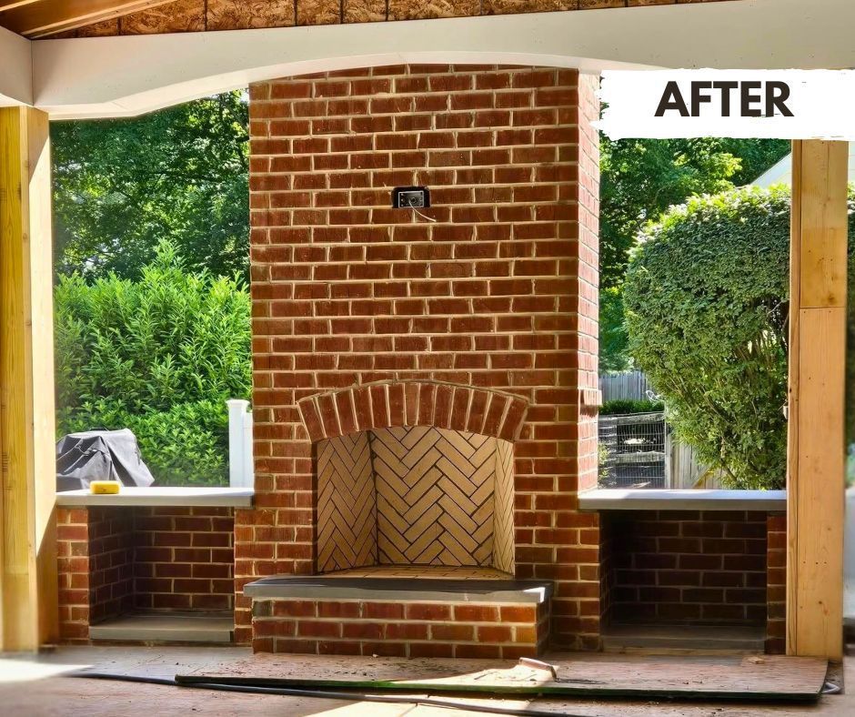 Outdoor brick fireplace with built-in shelves, featuring a herringbone brick firebox, set against a backdrop of lush greenery.