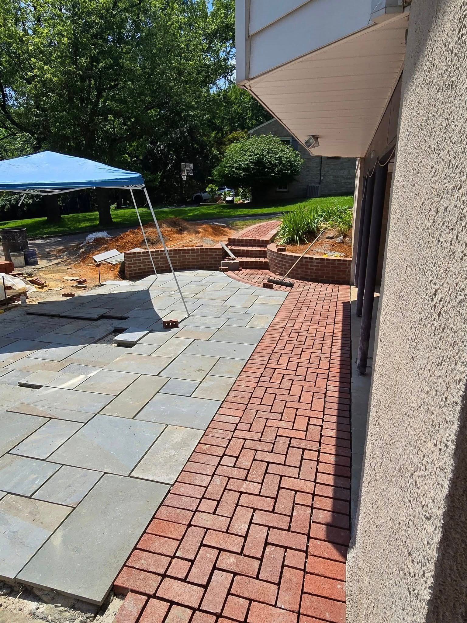 A patio under construction. Gray stone pavers and red brick border the side of a building. A blue tent is in the background.