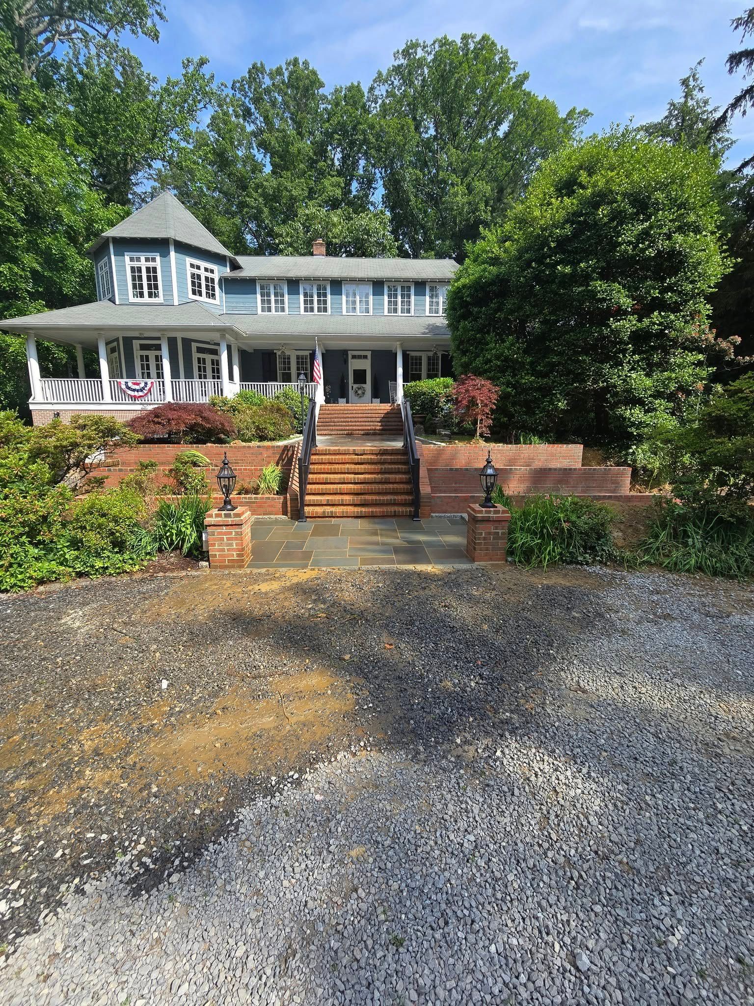 Blue house with a wraparound porch and brick steps leading up from a gravel driveway. Trees frame the house.
