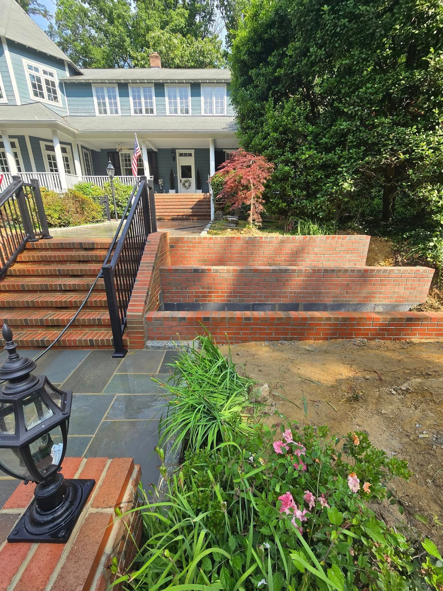 A multi-level brick and stone walkway leads to a light blue house with a porch. Green foliage frames the walkway.