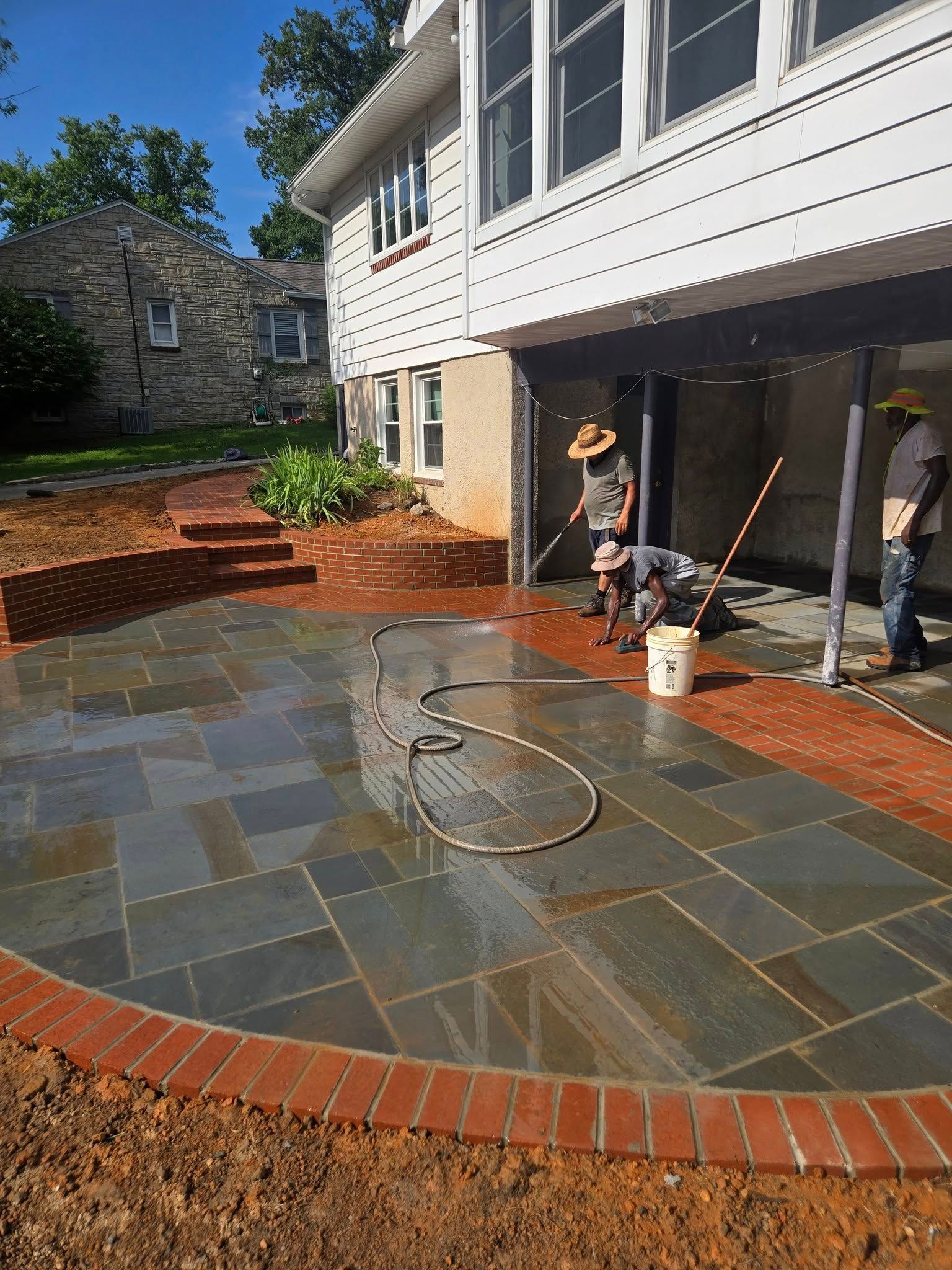 Construction of a patio with blue-gray flagstones and a brick border. Workers are present near a house.
