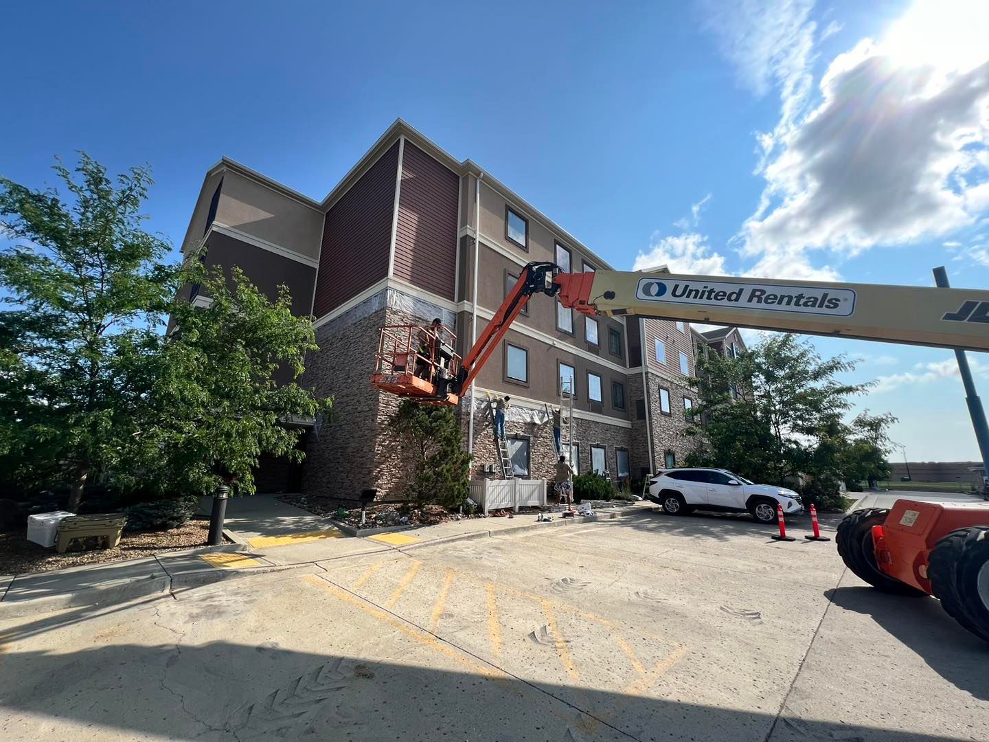 Construction work on a multi-story building. An aerial lift is used to access the exterior wall, with a white SUV and traffic cones in the parking lot.