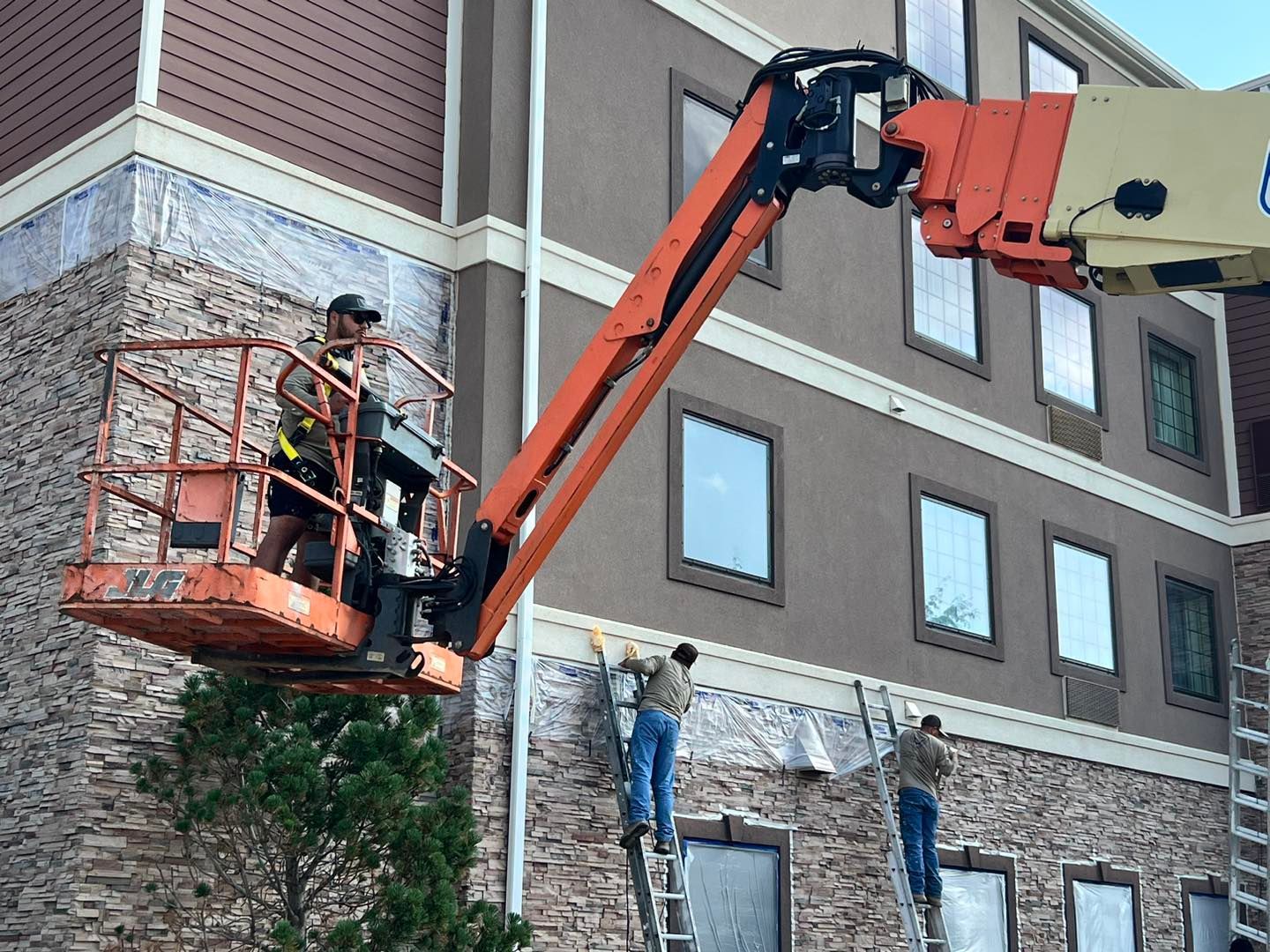 Construction workers applying material to a building's exterior, using a lift and ladders.