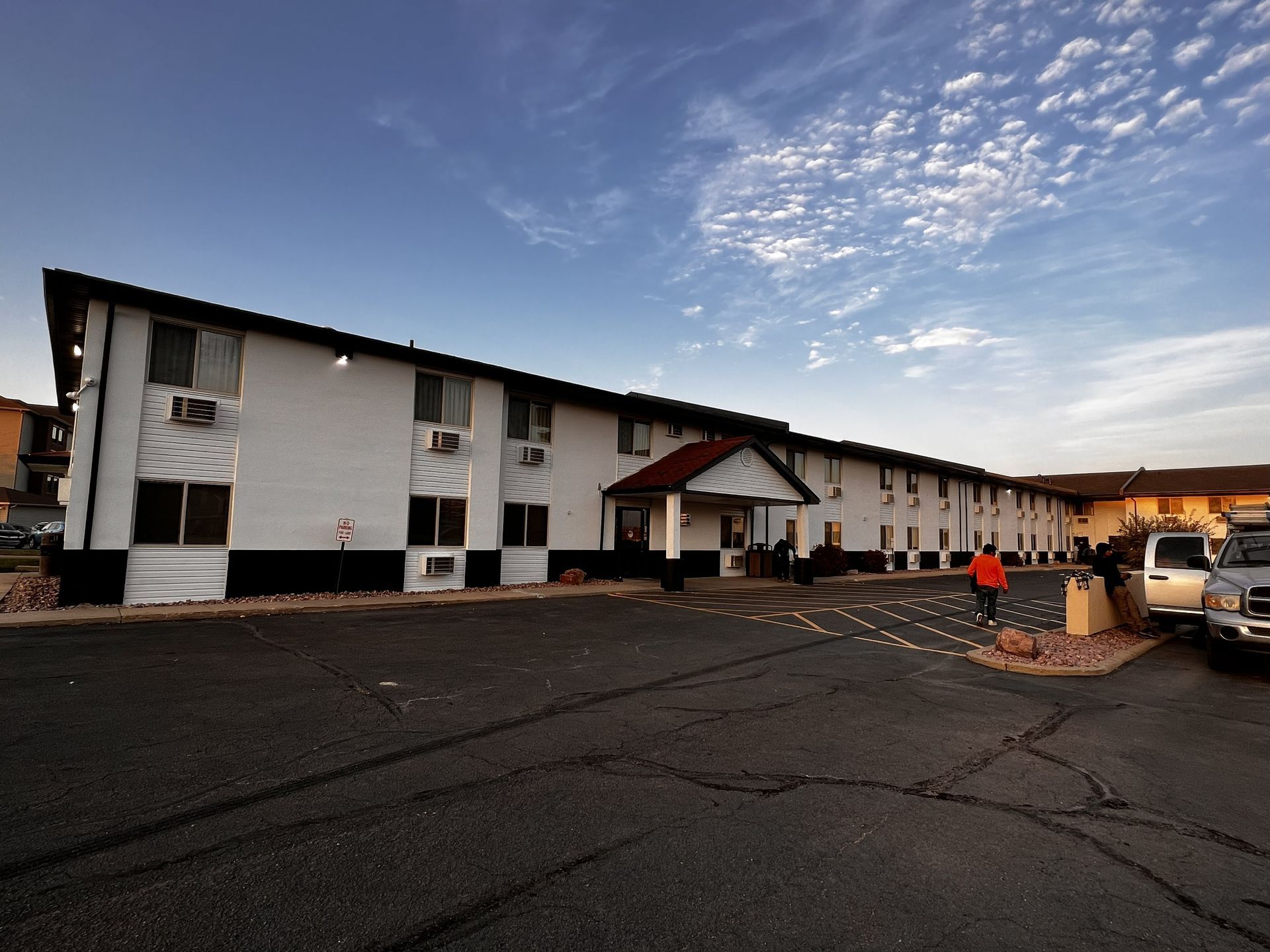 Two-story motel, white with black trim. Parked cars, asphalt parking lot, blue sky. A person walks.