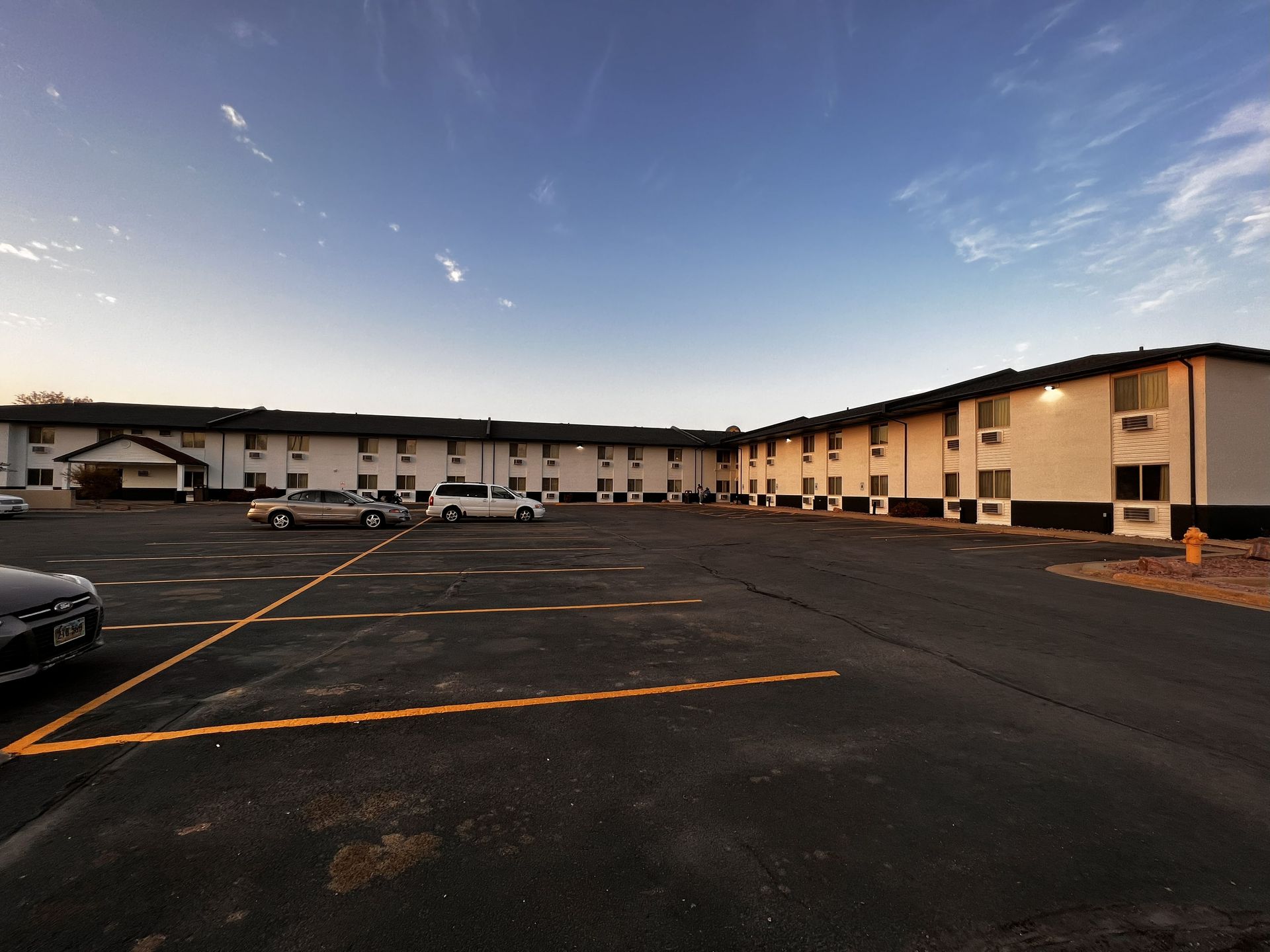 Exterior view of a motel building with a large asphalt parking lot. Cars parked, blue sky overhead.