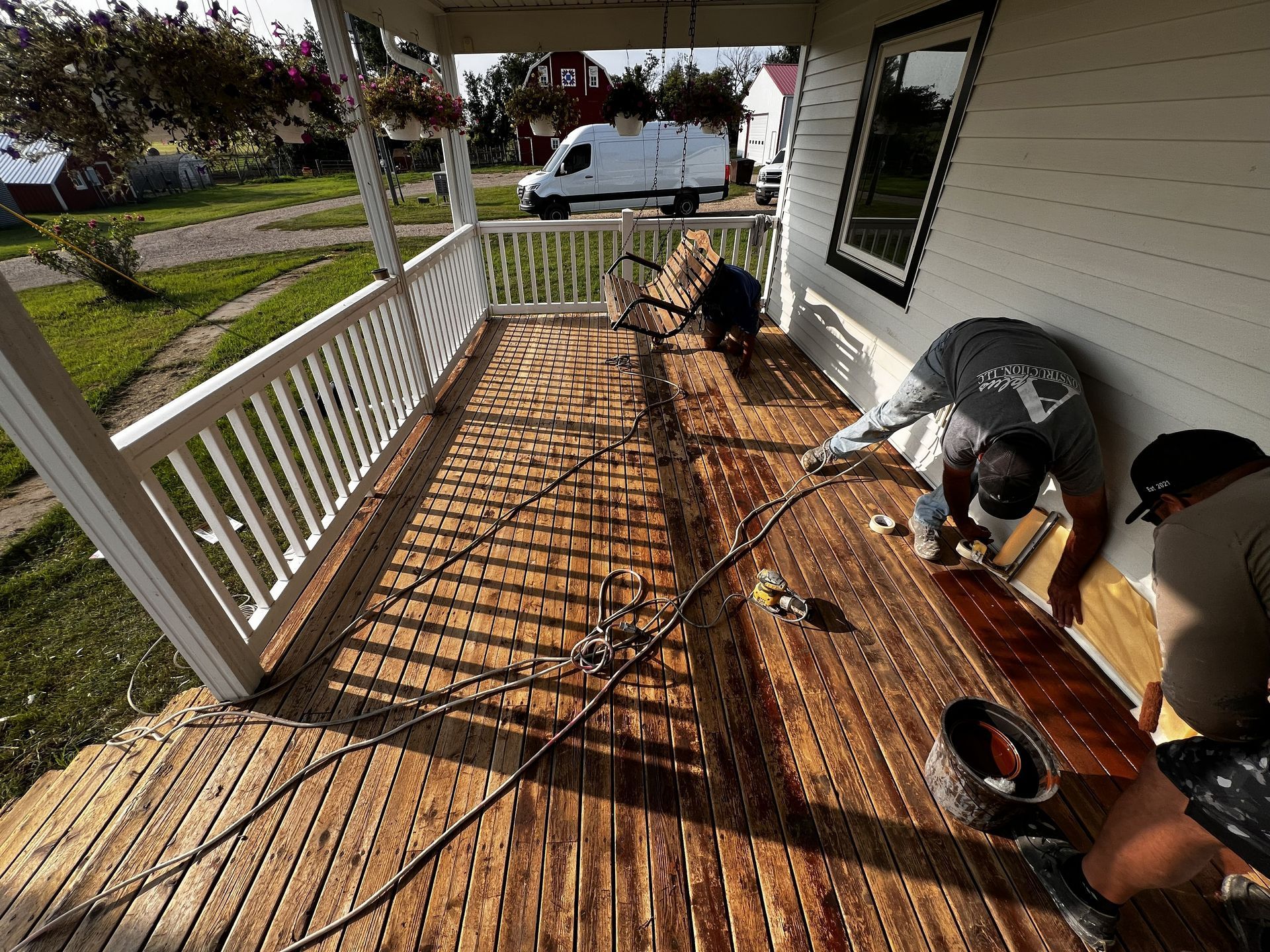 Men installing wooden planks on a porch. Sunlight casts shadows. White house, greenery, and a van are visible.