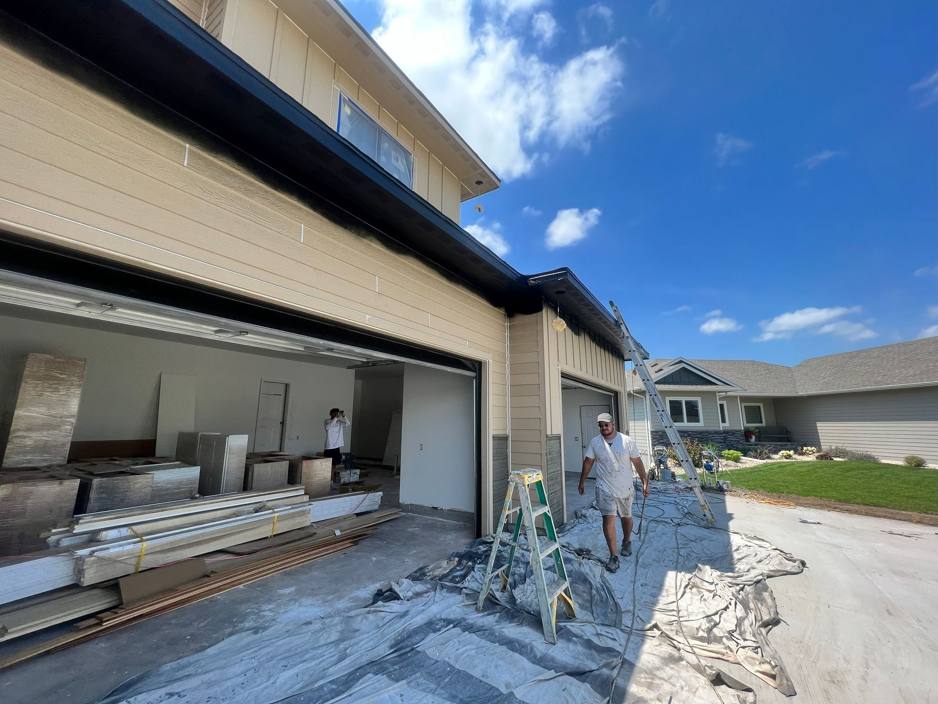 Exterior house under construction; two workers in garage, one by ladder. Light tan siding, blue sky.