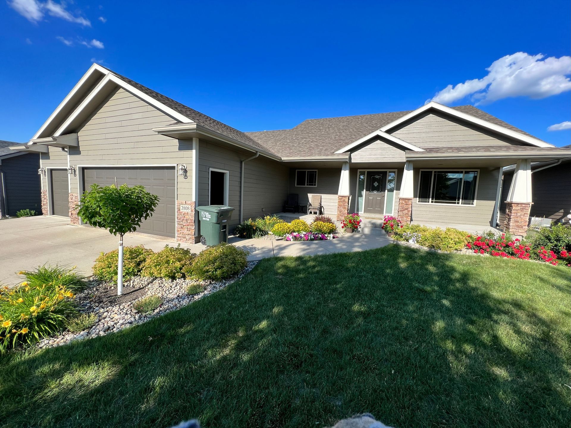 A one-story house with a green lawn, garden beds, and a driveway on a sunny day.