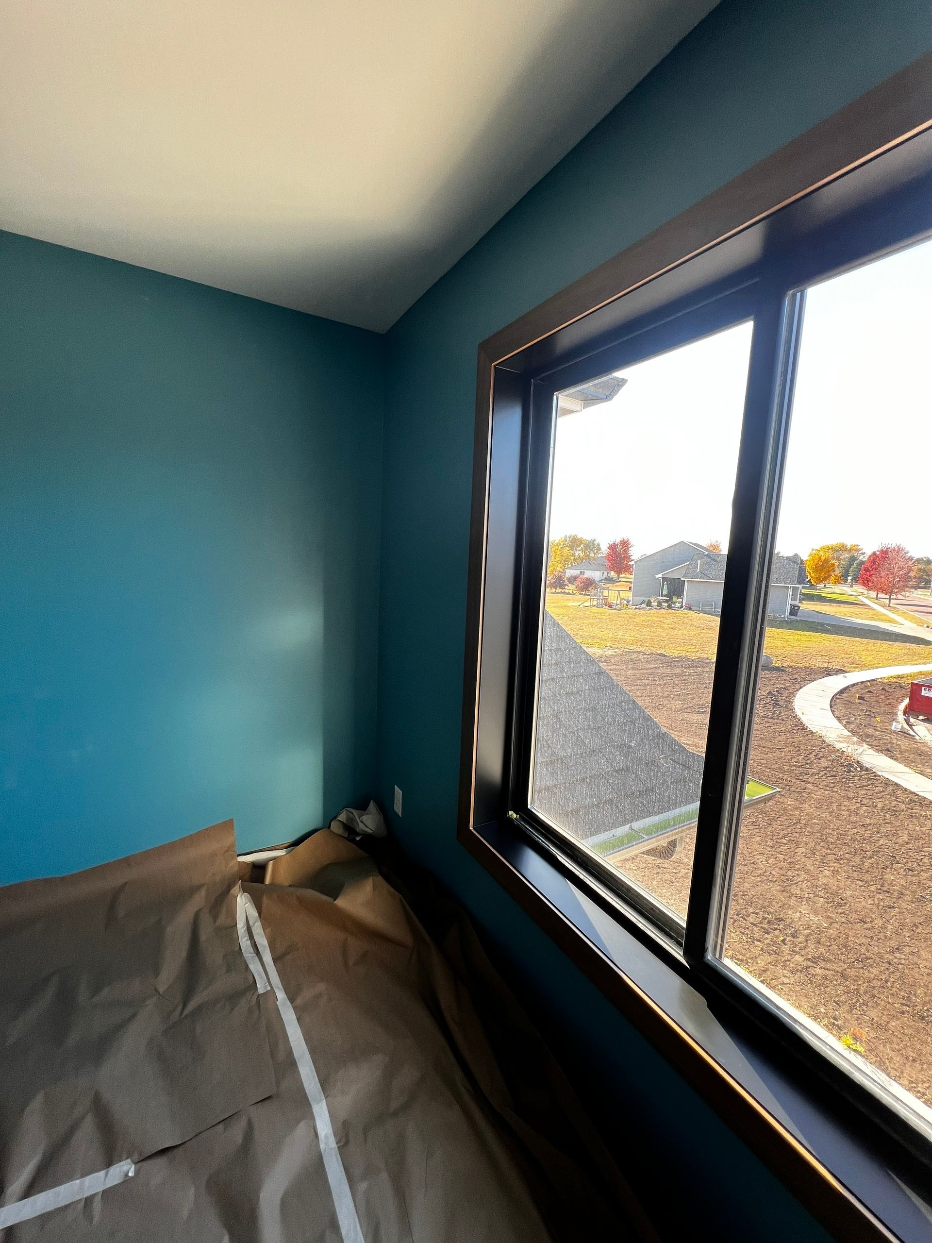 Interior corner with teal wall, dark-framed window. Brown paper protects a surface; a sunny outdoor view.