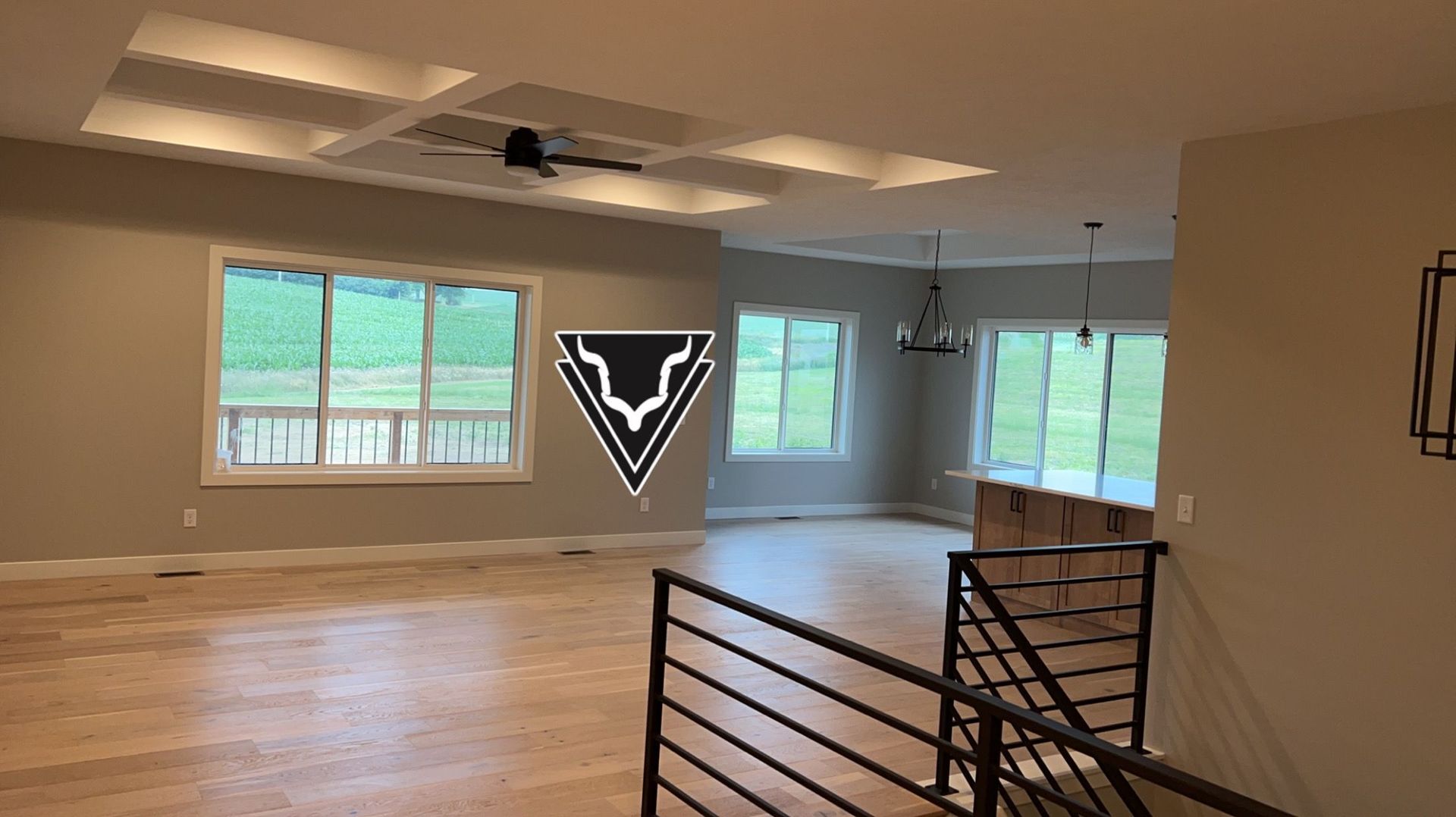 Open-concept interior with wood floors, large windows, and coffered ceiling. A staircase with black railing is in the foreground.
