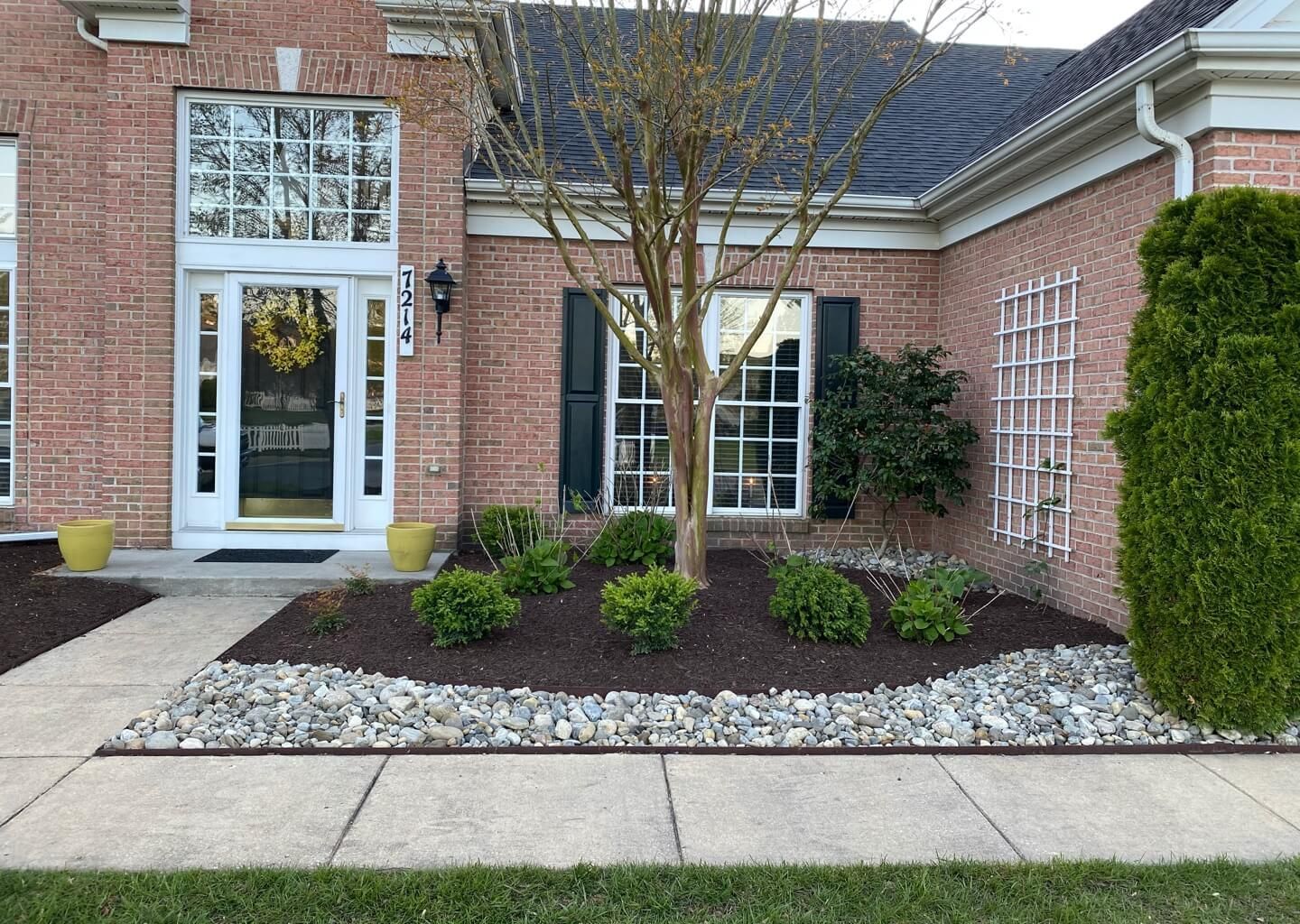 Brick house entry with white door, bay window, and landscaped front bed with small shrubs and rocks
