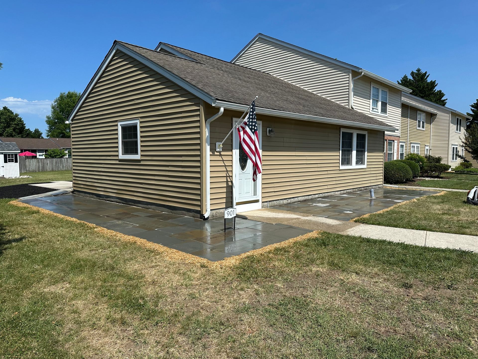 Single-story tan house with an American flag and wet concrete walkway in a suburban neighborhood