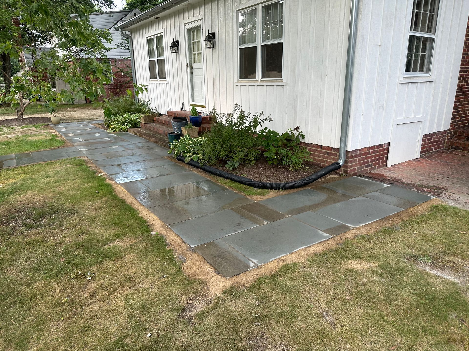 Wet concrete patio beside a white house, with a flower bed and wet lawn