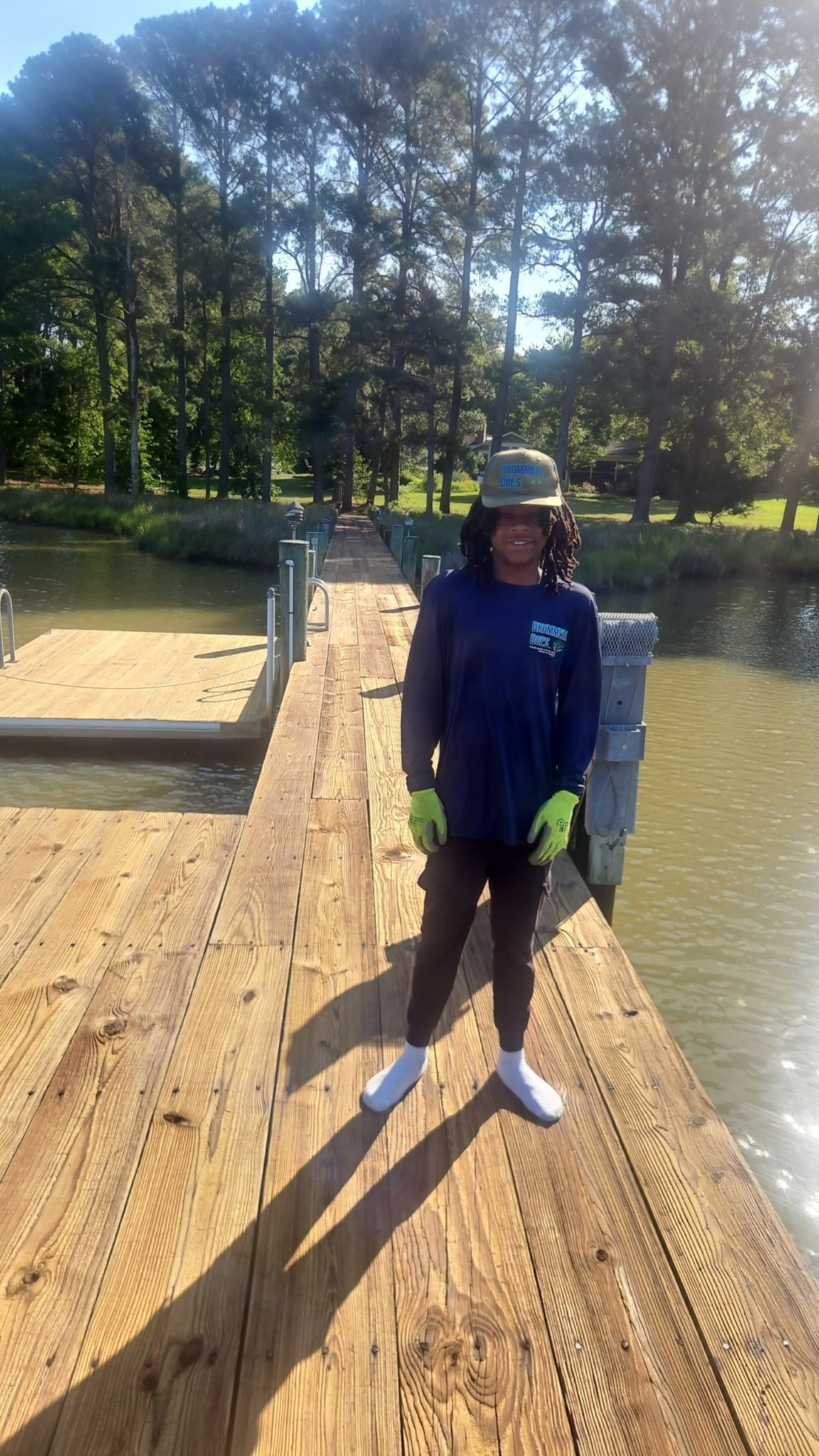 Person standing on a wooden dock by a calm lake, wearing a blue jacket and cap, with trees in the background