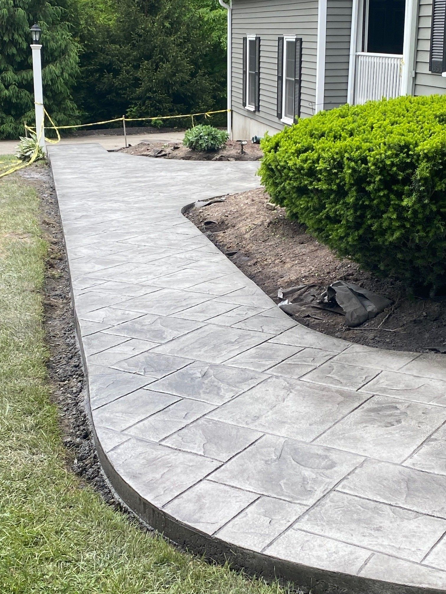 A gray, patterned concrete walkway curves past a residential house exterior with a large green shrub.
