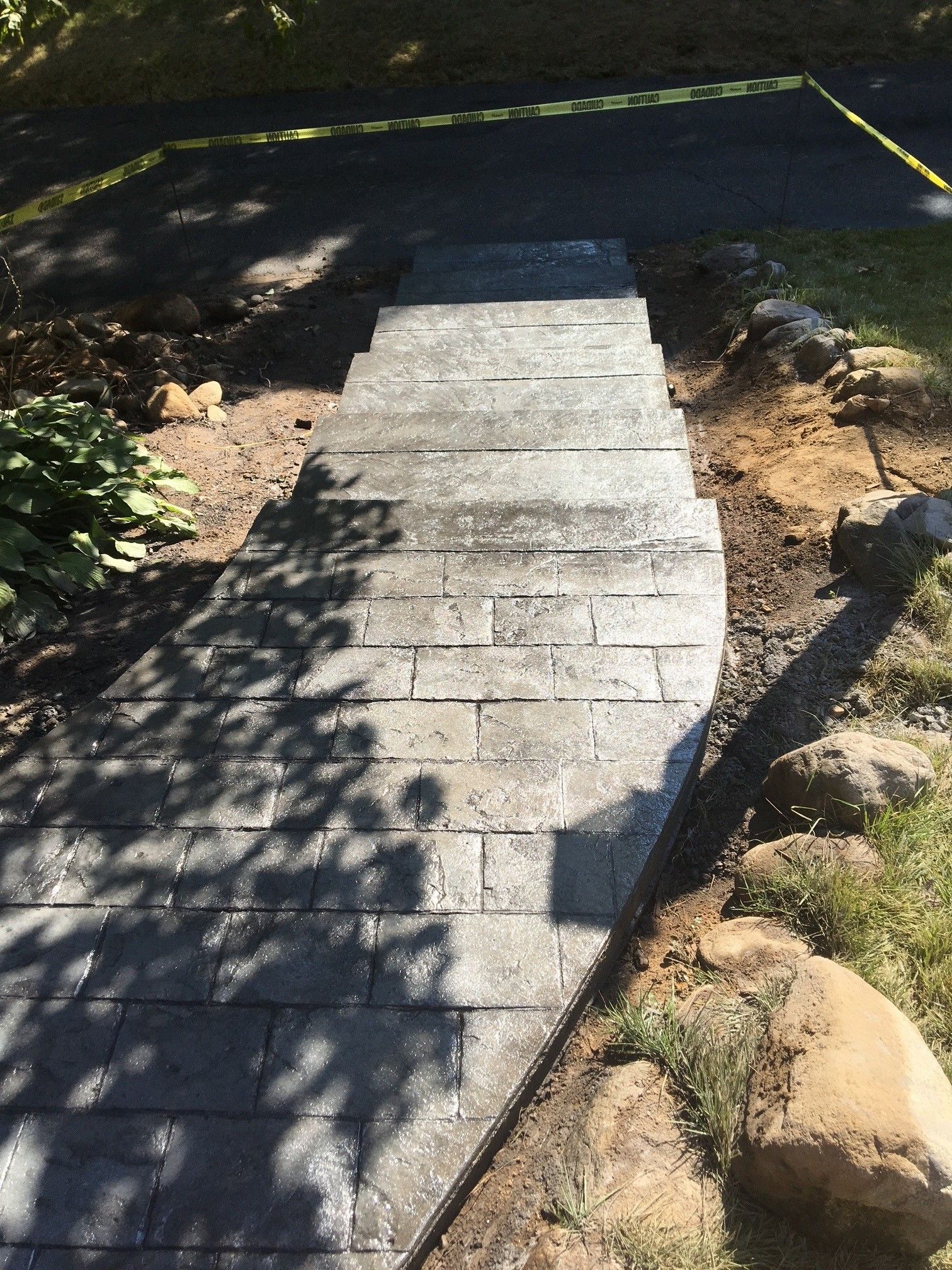 A stone paver walkway with steps leading down from a dark asphalt surface, flanked by patches of dirt and large rocks.