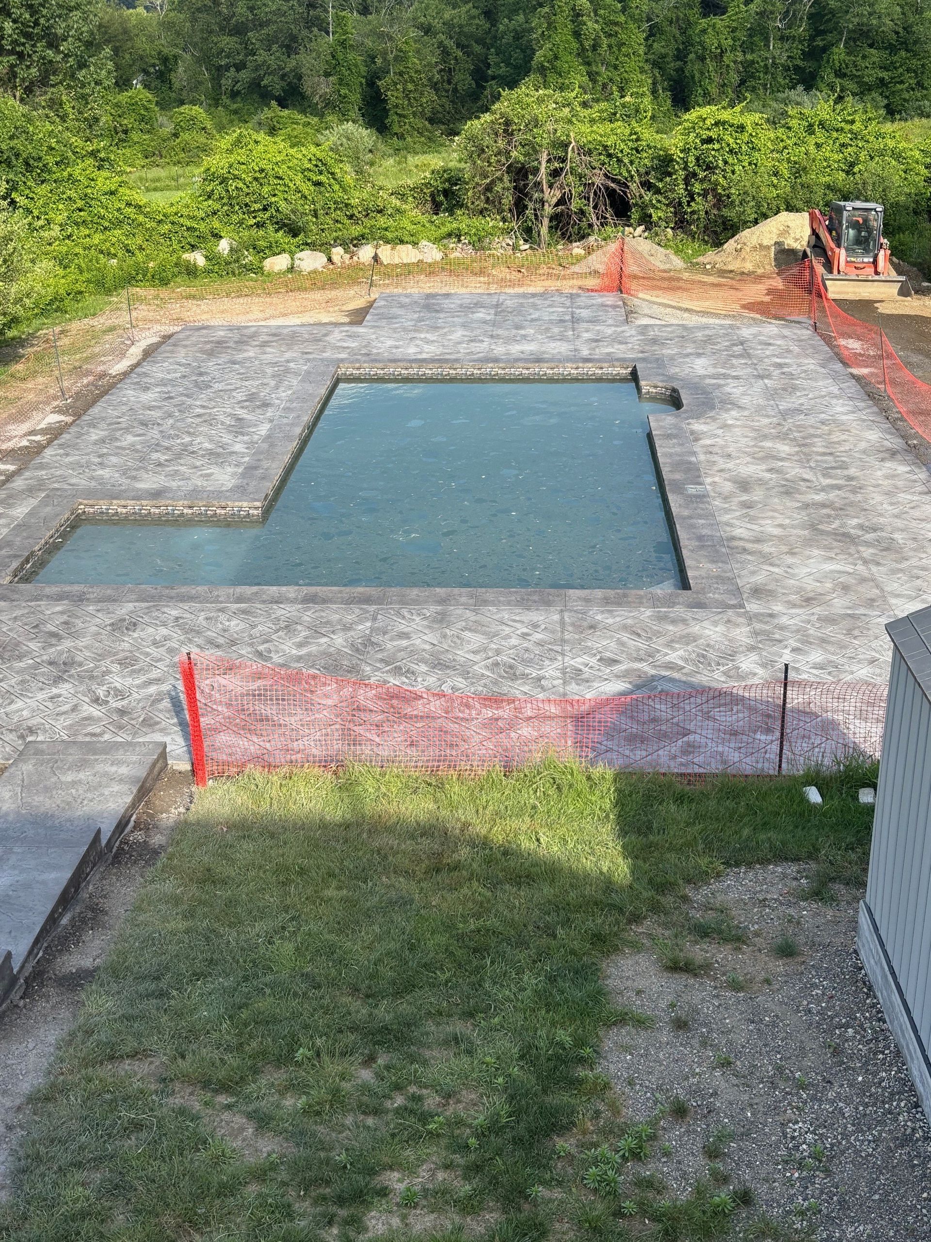 An L-shaped swimming pool under construction, surrounded by gray stone pavers, bordered by orange safety netting.