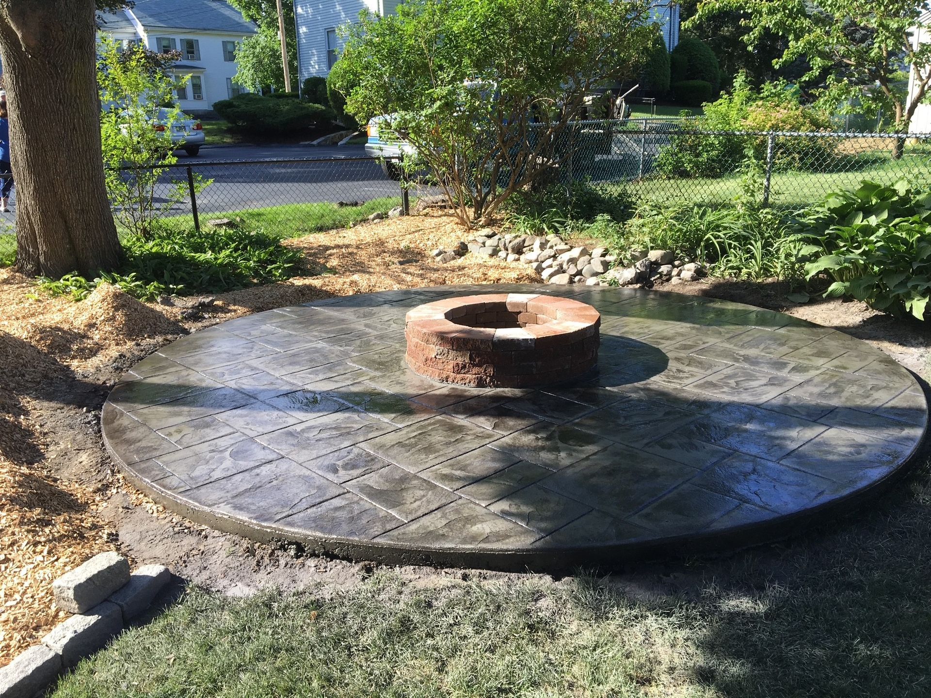 A circular, slate-gray stamped concrete patio featuring a central brick fire pit, situated in a backyard setting.