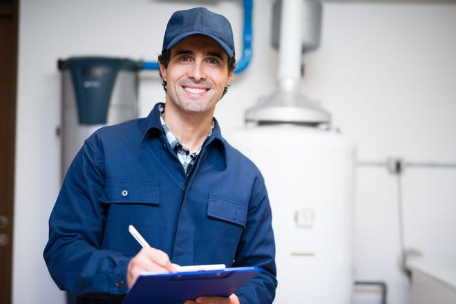 Technician in blue uniform and cap smiling while writing on a clipboard in a utility room