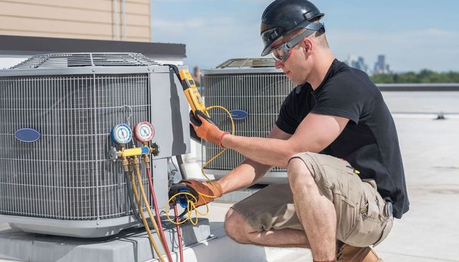 Technician kneeling beside rooftop HVAC unit, inspecting gauges and wiring under a hard hat.