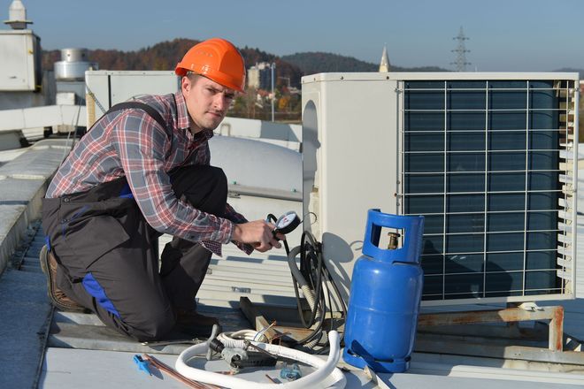 Worker in orange hard hat servicing rooftop HVAC equipment beside a blue gas cylinder.