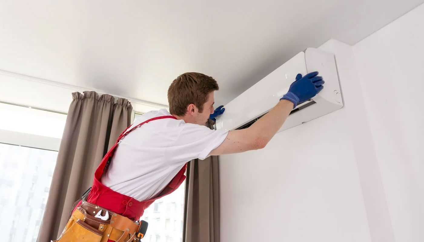 Technician cleaning a wall-mounted air conditioner with a blue cloth in a bright room