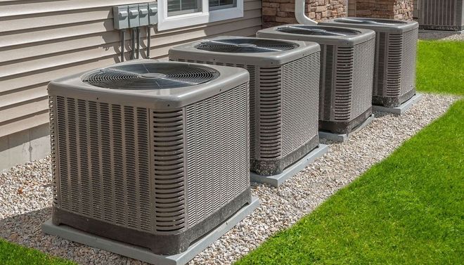 Row of outdoor air conditioning units beside a house, on a gravel strip next to green lawn.