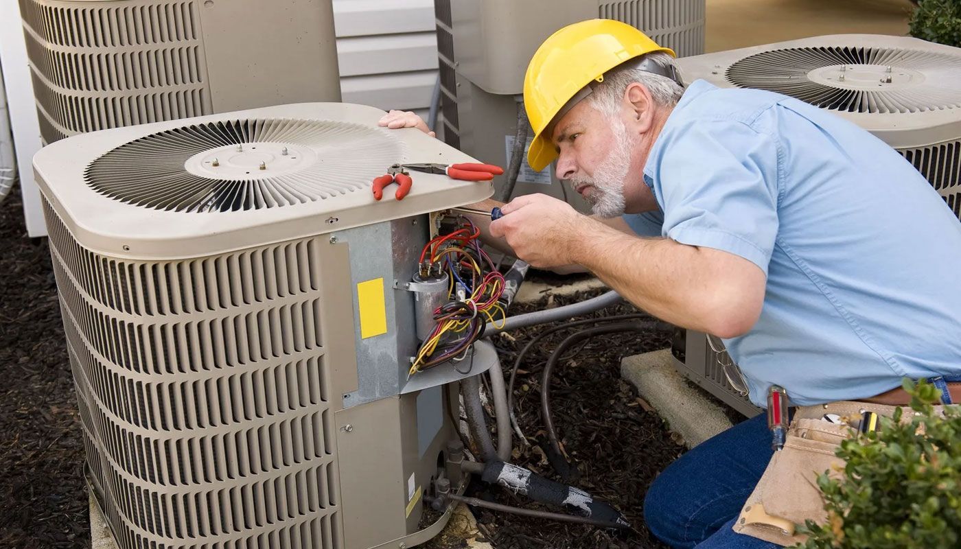 Technician in a yellow hard hat repairs outdoor HVAC unit beside a building.