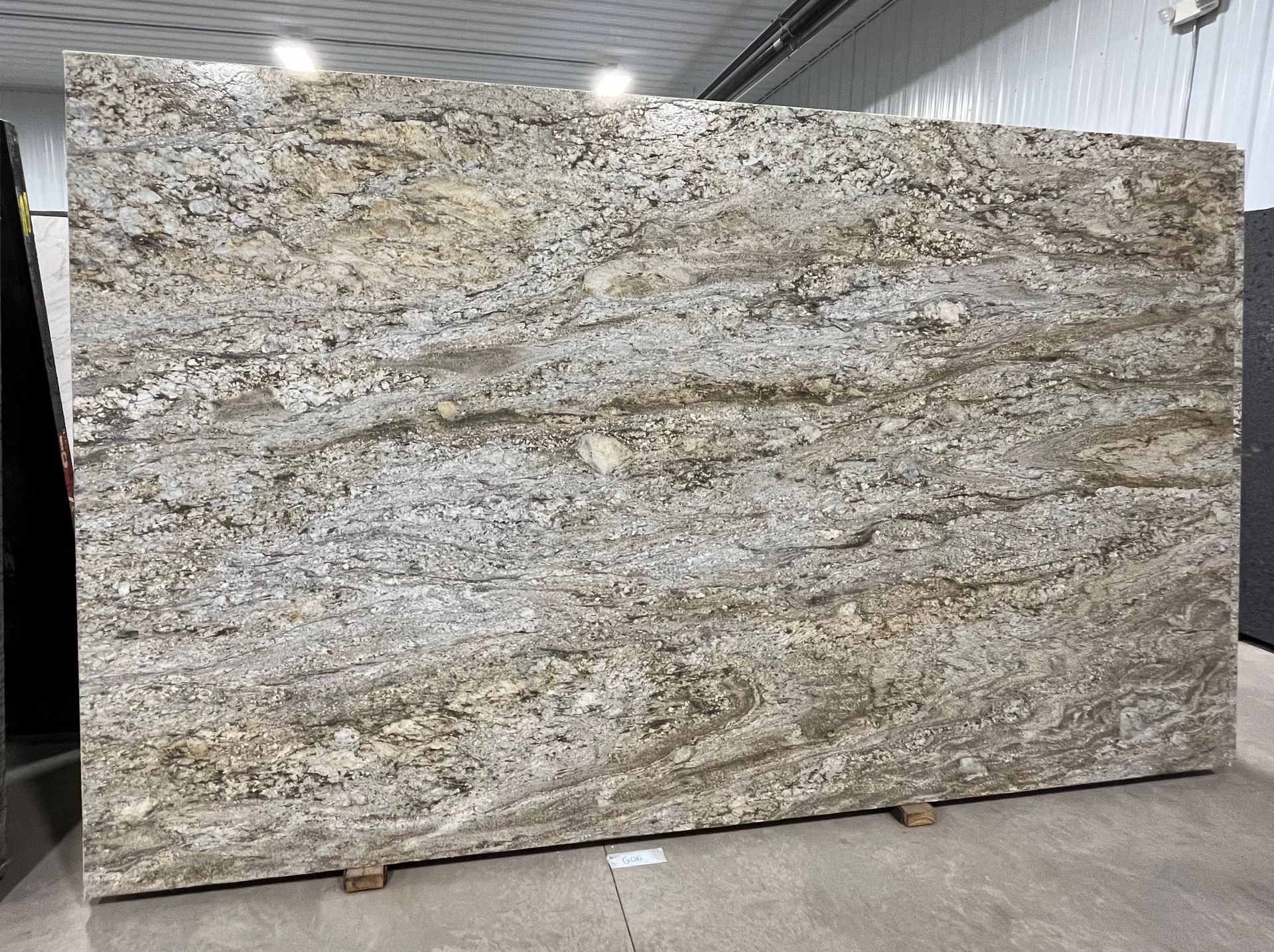 Large polished granite slab with beige, gray, and gold veining displayed in a warehouse.