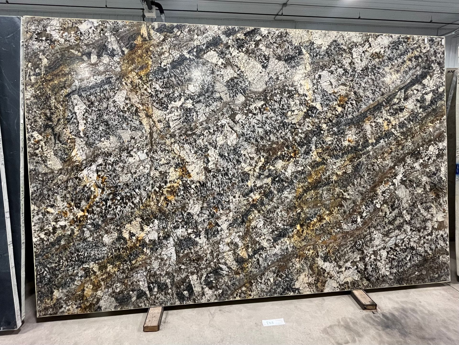 Large polished granite slab with gray, white, and gold veining displayed upright in a warehouse.