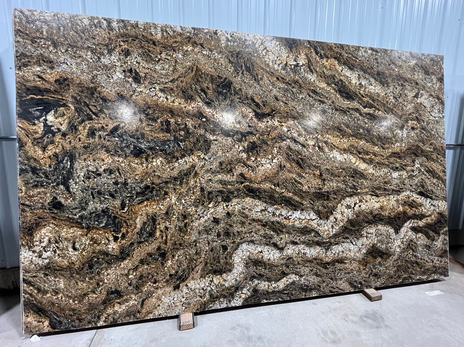 Large polished granite slab with brown, black, and cream wavy patterns leaning outdoors