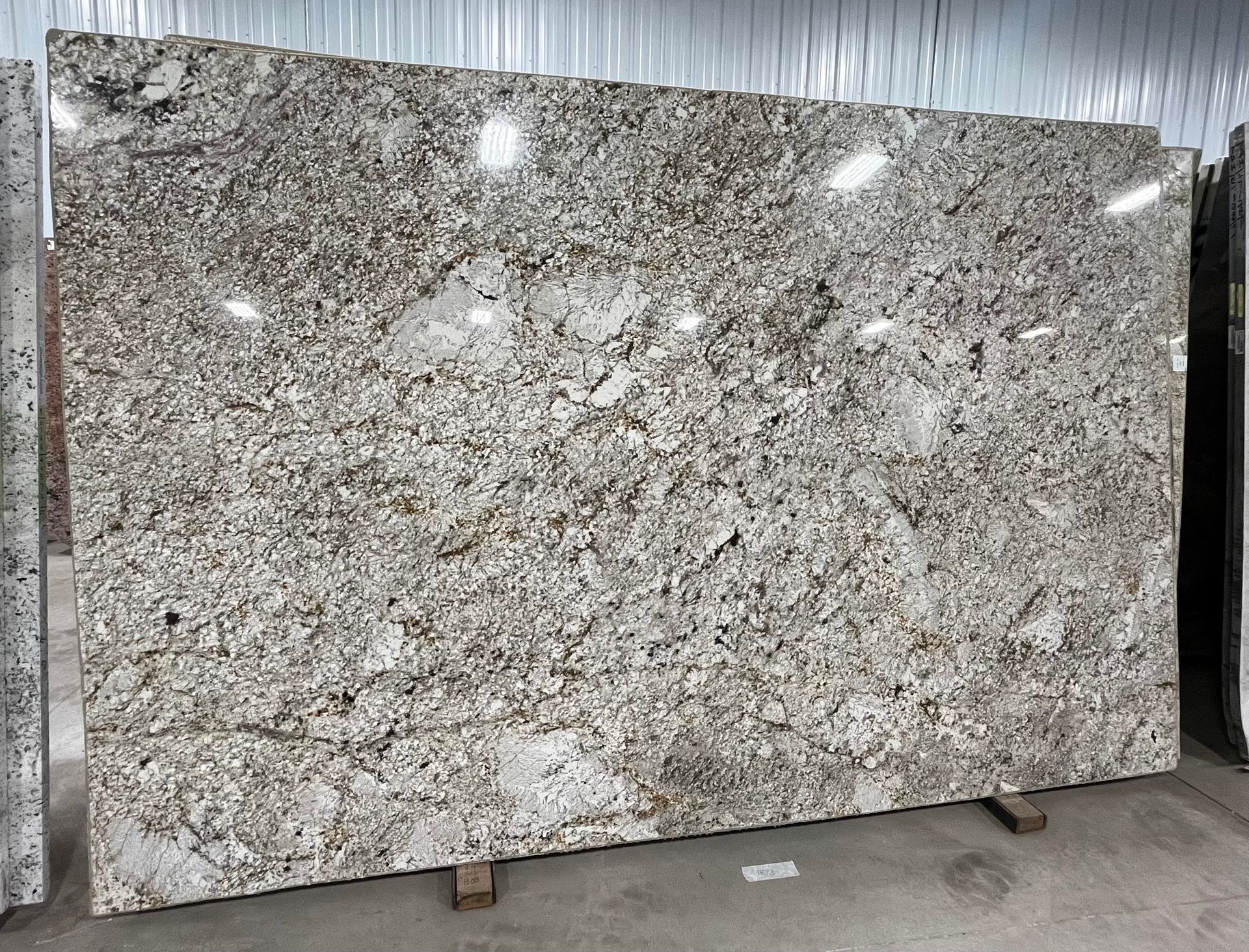 Large polished granite slab with white, gray, and black speckles displayed upright in a warehouse.