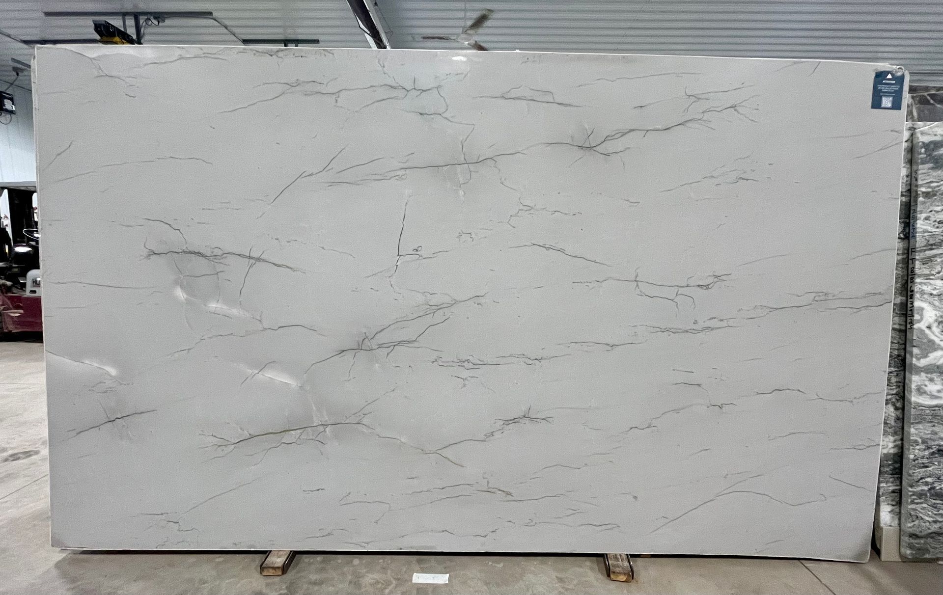 A large slab of light grey quartz stone with subtle, thin dark grey veining, displayed vertically in a warehouse setting.