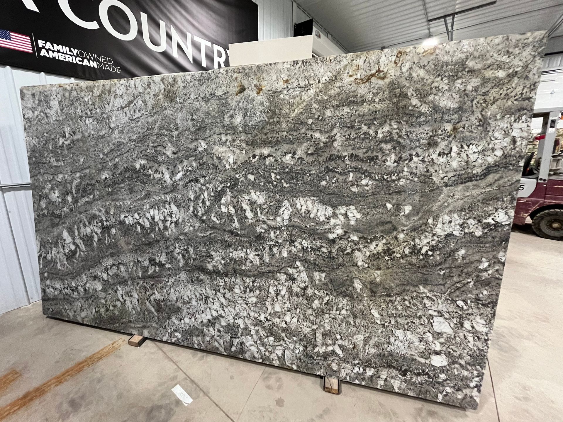 A large, polished slab of granite featuring swirls of charcoal gray, white, and earthy brown tones in a warehouse setting.